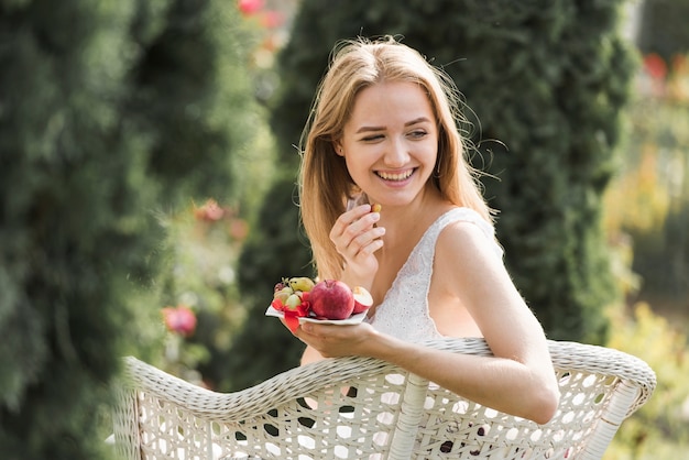 Souriante blonde jeune femme assise sur une chaise en mangeant des fruits dans le jardin Photo gratuit