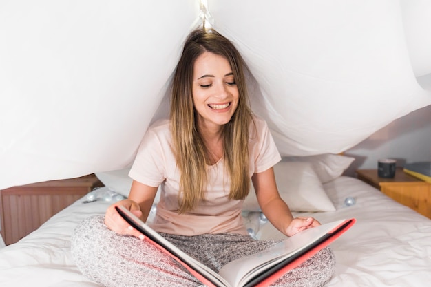 Souriante Blonde Jeune Femme Assise Sur Le Lit En Lisant Le Magazine