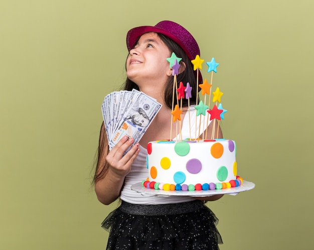 Souriante Jeune Fille Caucasienne Avec Chapeau De Fete Violet Tenant Un Gateau D Anniversaire Et De L Argent Regardant Le Cote Isole Sur Un Mur Vert Olive Avec Espace De Copie Photo Gratuite