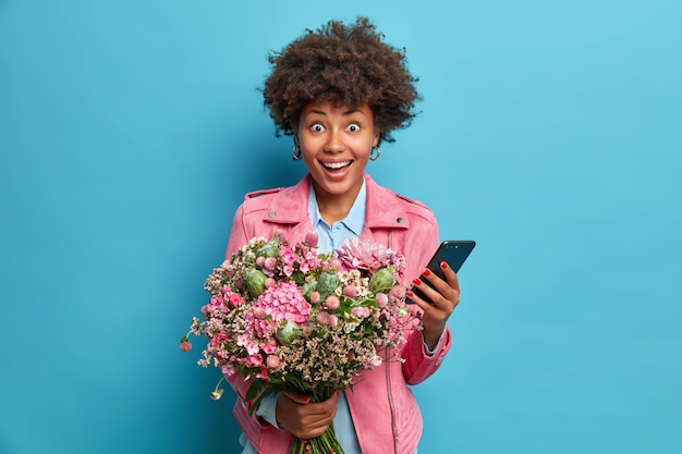 Surpris Femme Gaie Tient Le Telephone Portable Moderne Accepte Les Felicitations Pour Son Anniversaire Pose Avec Bouquet De Fleurs De Fete Isole Sur Mur Bleu Photo Premium