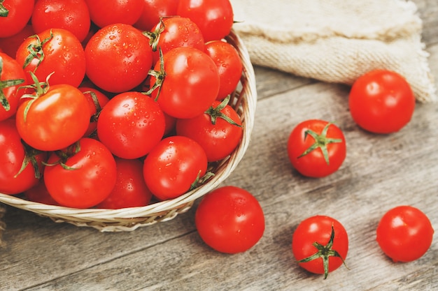 Tomates rouges fraîches dans un panier en osier sur une vieille table en bois. tomates cerises mûres et juteuses avec des gouttes d'humidité, table en bois gris, autour d'un chiffon de toile de jute. dans un style rustique. Photo Premium