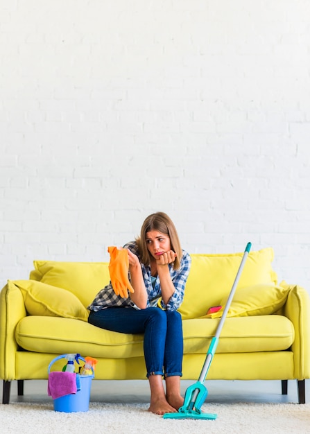 Triste jeune femme assise sur un canapé jaune regardant des gants de caoutchouc orange Photo gratuit
