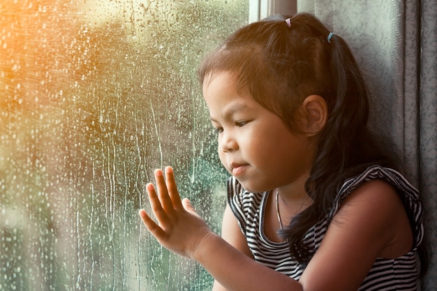 Triste Petite Fille Asiatique Regardant à Travers La Fenêtre Dans La