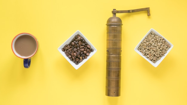Vieux moulin à café; grains de café crus et torréfiés et tasse à café sur fond jaune vif Photo gratuit