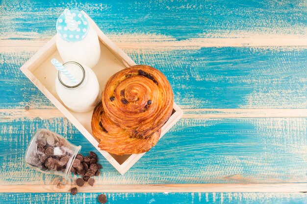 Vue d'angle élevé de brioches à la cannelle et de bouteilles de lait dans un plateau en bois près de chips de choco renversées dans un bocal en verre Photo gratuit