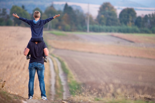 Vue arrière du père sportif portant sur les épaules fils marchant à travers un terrain herbeux sur les arbres vert brumeux floues et ciel bleu. Photo Premium