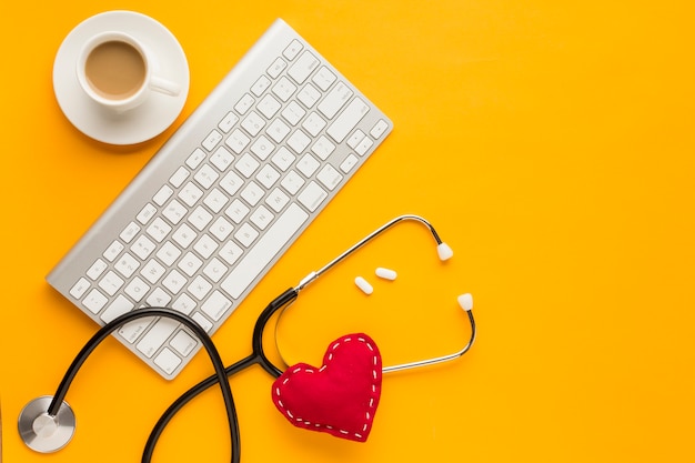 Vue de dessus du clavier sans fil; comprimés; tasse à café; stéthoscope; coeur de jouet cousu; au-dessus de la toile de fond jaune Photo gratuit