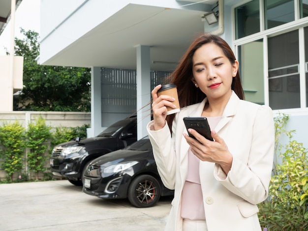 Aziatische de koffiekop van de vrouwenholding en het gebruiken van smartphone terwijl status voor huis. Premium Foto