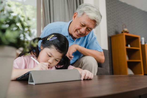 Aziatische grootvader leert kleindochter thuis tekenen en huiswerk maken. hogere chinees, gelukkige opa ontspant met jong meisje liggend op bank in woonkamer thuis concept. Gratis Foto