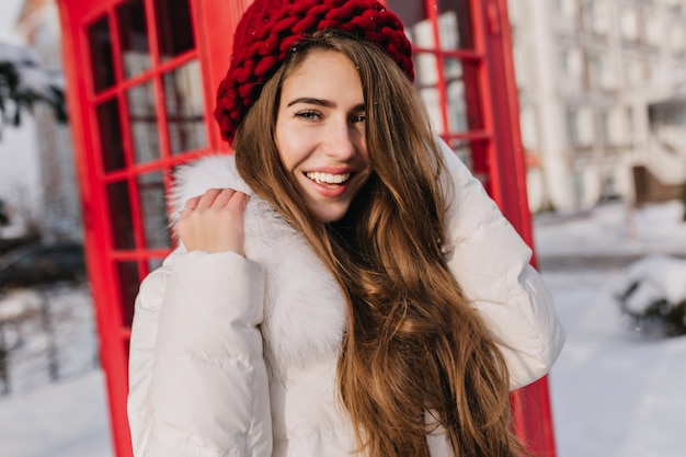 Close Up Portret Van Gelukkige Vrouw Met Glanzend Bruin Haar Poseren Naast Rode Telefooncel Buitenfoto Van Een Prachtig Vrouwelijk Model In Gebreide Baret Die Geniet Van Een Ijzige Ochtend In Engeland Gratis