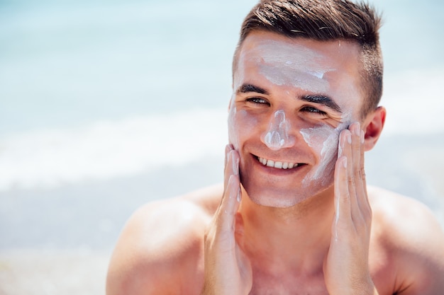 De glimlachende mens die het looien room op zijn gezicht zet, neemt een sunbath op het strand. Gratis Foto
