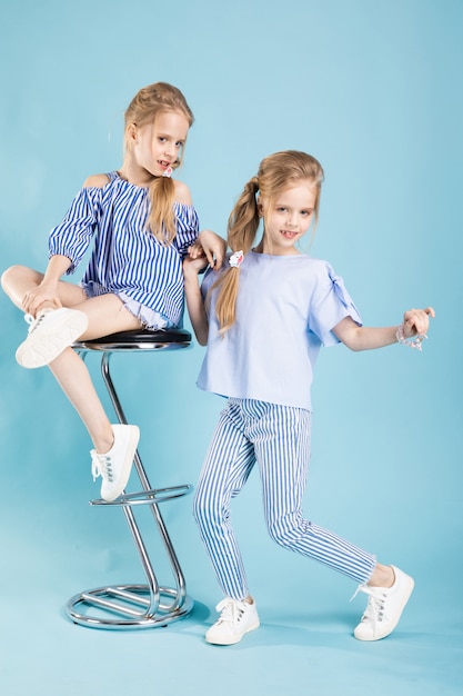 De meisjestweelingen in lichtblauwe kleren stellen dichtbij een barkruk op een blauw. Premium Foto