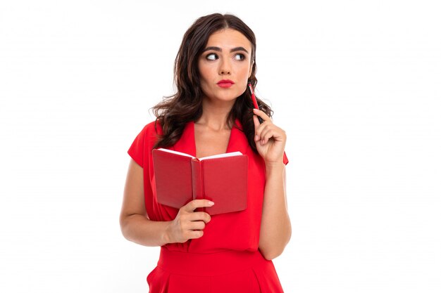 Een jonge vrouw met rode lippen, lichte make-up, een oogverblindende glimlach, donker golvend lang haar, in een rode zomerjurk, houdt een notitieboekje vast en denkt na Premium Foto