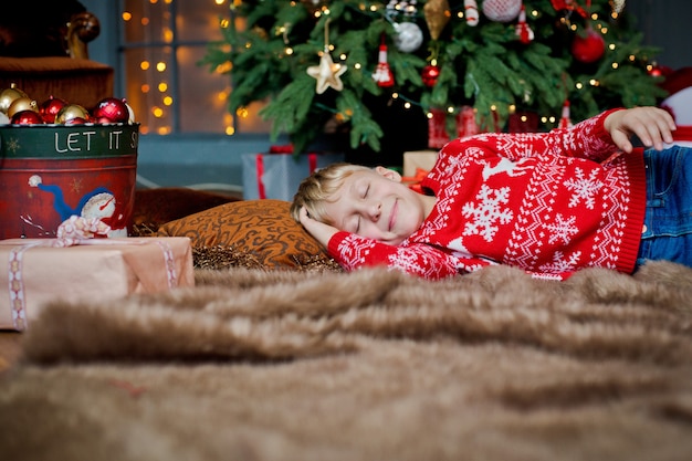 Een kind slaapt op kerstavond onder een versierde kerstboom en wacht op een geschenk. familie viert kerstmis thuis. de kinderen slapen. Premium Foto
