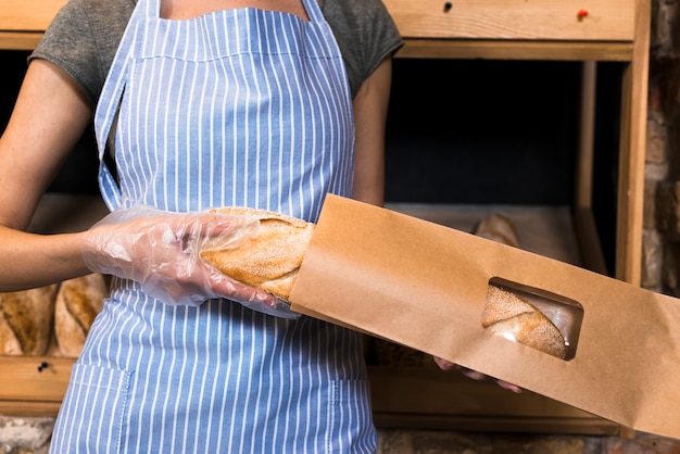 Een vrouwelijke bakker in schort die het baguette brood in de bruine document zak inpakken Gratis Foto