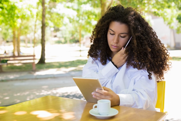 Geconcentreerde dame die telefoon uitnodigt en tablet in openluchtkoffie gebruikt Gratis Foto