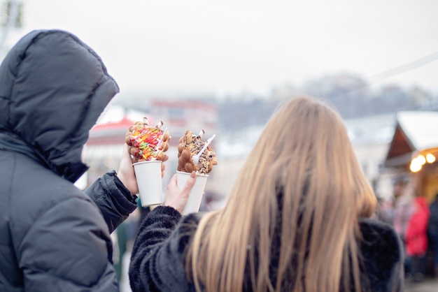 Gelukkig paar dat in warme kleren in liefde bellenwafels eet bij kerstmismarkt. vakanties, winter, kerstmis en mensen Premium Foto