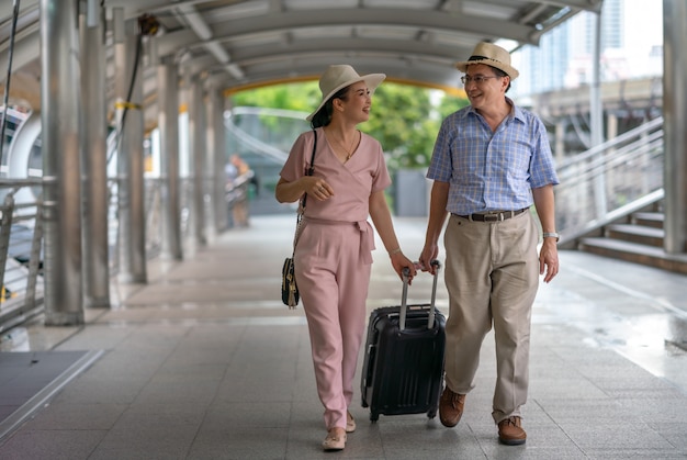 Gelukkige aziatische paar hogere toeristen die kofferhandvat in de stad houden terwijl het reizen Premium Foto