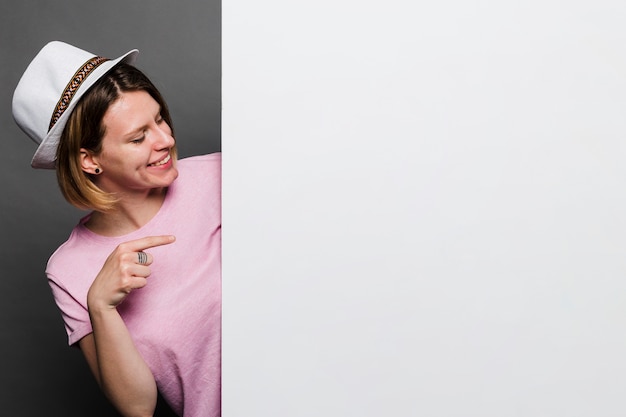 Glimlachende jonge vrouw die witte hoed draagt die vinger richt naar witte kaart Gratis Foto