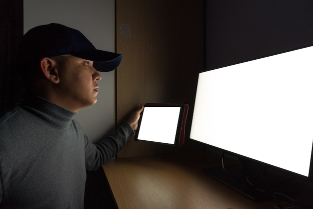 Het zijaanzicht van mensenhakker zit bij de computermonitor, witte het schermtablet in de donkere ruimte. Premium Foto
