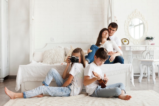 Houdende van ouderzitting op bed en hun camera van de dochterholding en zoon die ukelele spelen Gratis Foto