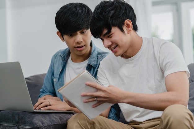 Jonge aziatische homo paar werkende laptop bij modern huis. azië lgbtq + mannen gelukkig ontspannen plezier met behulp van computer en het analyseren van hun financiën op internet samen liggend bank in de woonkamer in huis. Gratis Foto