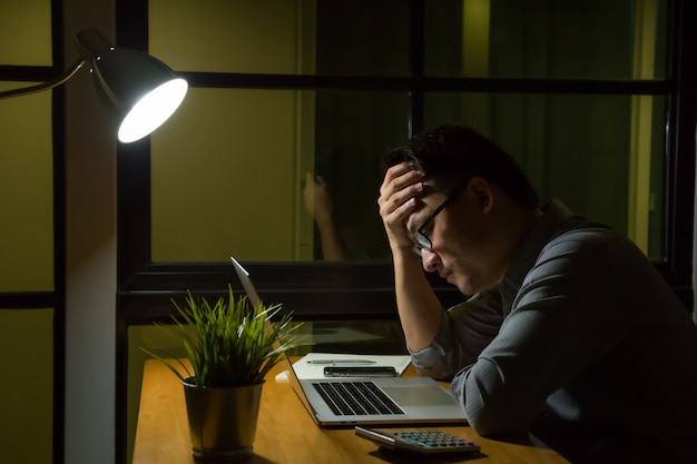 Jonge aziatische mensenzitting op bureaulijst die laptop computer in het donkere recente nacht werken werken die het ernstige denken en spanning op kantoor werken. overuren en hard werken concept Premium Foto