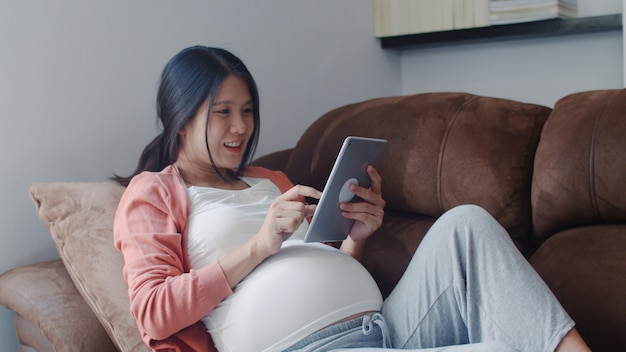 Jonge aziatische zwangere vrouw die de zwangerschapsinformatie van het tabletonderzoek gebruikt. mamma die het gelukkige positief en vreedzame glimlachen voelen terwijl zorg haar kind thuis liggend op bank in woonkamer. Gratis Foto