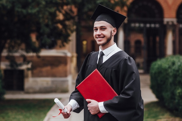 Jonge knappe afgestudeerde man in mantel bedrijf mate in de buurt van de universiteit Premium Foto
