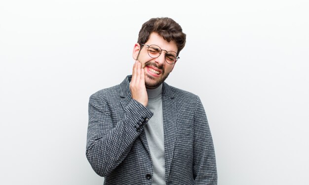 Jonge manager man met wang en pijnlijke kiespijn, ziek, ellendig en ongelukkig, op zoek naar een tandarts witte muur Premium Foto