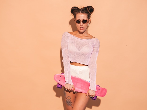 Jonge mooie sexy lachende hipster vrouw in zonnebril. trendy meisje in zomer gebreid vest onderwerp, shorts. positieve vrouw gek met roze cent skateboard, geïsoleerd op beige muur. twee hor Gratis Foto