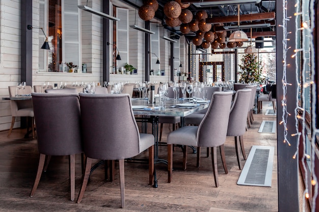 Kerstverlichting, decor, interieur modern restaurant, panoramische ramen, instelling, serveren banket, grijze textiel stoelen, serveertafels, wijnglas, borden, bestek. feestelijk nieuwjaar, winter Premium Foto