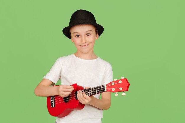Leuke jongen speelt de ukelele. gelukkig kind genieten van de muziek. student die ukeleles spelen. modieuze jongen in zomer hoed geïsoleerd op groene achtergrond. | Premium Foto