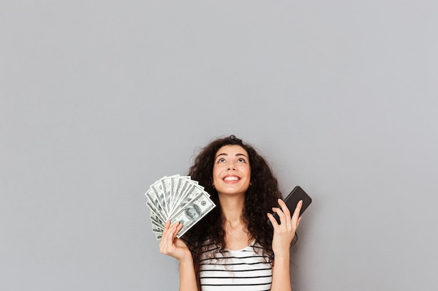 Leuke vrouw in gestreepte t-shirt met fan van 100-dollarbiljetten en mobiele telefoon in handen opzoeken die dankbaar is kan niet geloven in haar triomf Gratis Foto