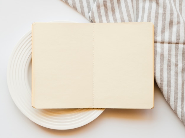 Lichtbruin schetsboek op een witte plaat op een witte achtergrond met een gestreept grijs en wit tafelkleed Gratis Foto