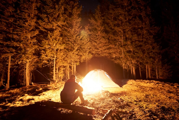 Mannelijke toerist rust 's nachts uit in zijn kamp bij het kampvuur en de tent onder een prachtige nachthemel vol sterren en de maan en geniet van een nachtscène in de bergen Premium Foto
