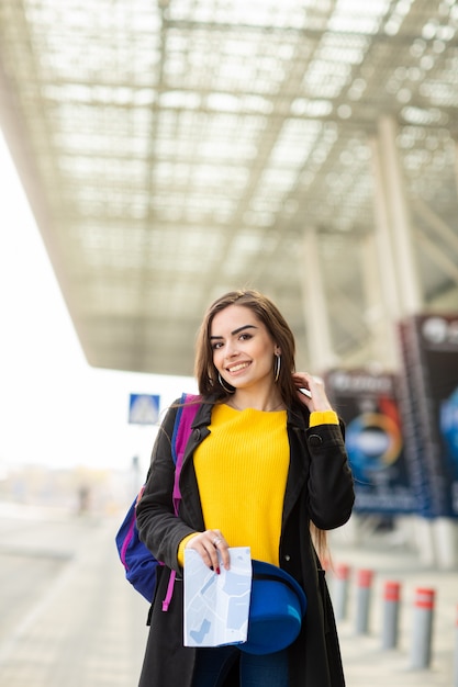 Mooi jong meisje met een rugzak achter haar schouder die een kaart, in de straat dichtbij de luchthaven houdt Premium Foto