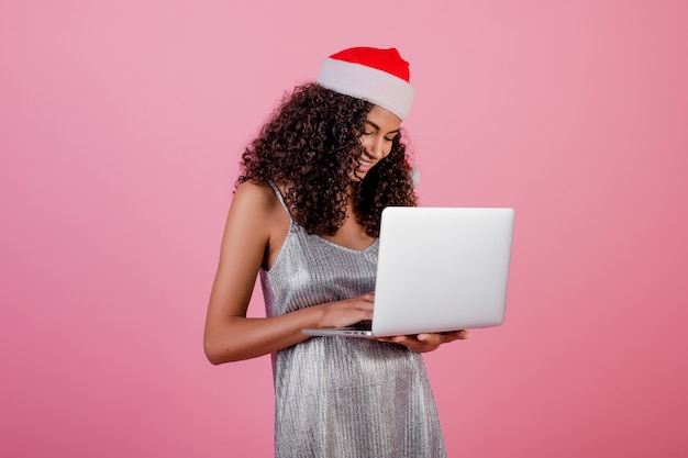 Mooi zwart meisje met laptop die de hoed en de kleding van vakantiekerstmis draagt die over roze wordt geïsoleerd Premium Foto