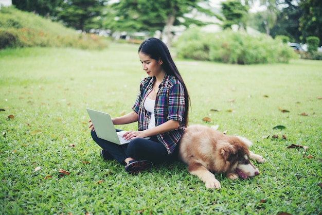 Mooie jonge vrouw die laptop met haar kleine hond in een park in openlucht met behulp van. levensstijl portret. Gratis Foto