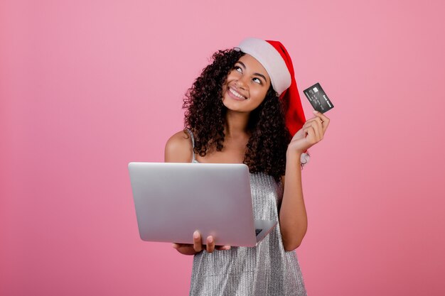 Mooie zwarte met creditcard en laptop die de hoed en de kleding van vakantiekerstmis dragen die over roze wordt geïsoleerd Premium Foto