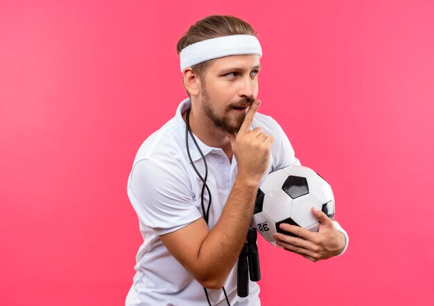 Overtollig spreken Rode datum Onder de indruk jonge knappe sportieve man met hoofdband en polsbandjes met  voetbal gebaren stilte en kijken naar kant met springtouw om zijn nek  geïsoleerd op roze muur | Gratis Foto