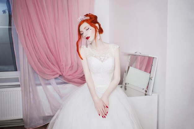 Roodharig meisje in een trouwjurk, helder ongewoon uiterlijk, rode nagels, een meisje met bleke huid, mooie trouwjurk, een hart op haar wang, lichte make-up, op een wit, fetisch model Premium Foto