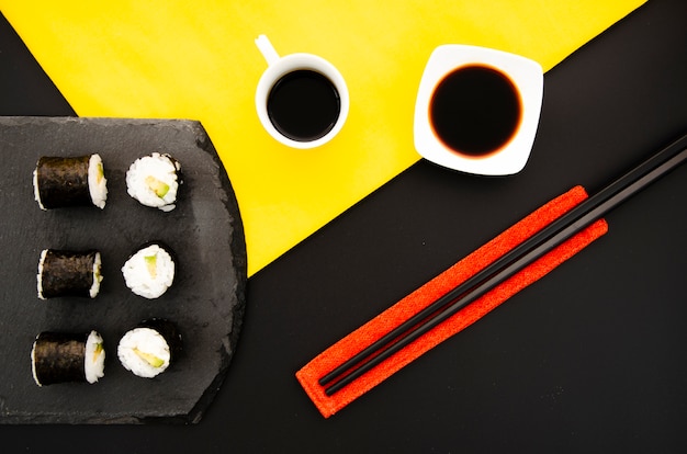 Steenplaat met sushibroodjes en kom met sojasaus op een zwarte achtergrond met eetstokjes Gratis Foto