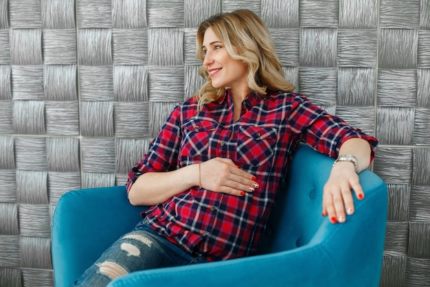 Stijlvolle aanstaande moeder vrouw teder omarmen en streelde haar buik. mooie blonde zwangere vrouw die als voorzitter in modieuze studio rust. gezonde zwangerschap en moederschap. Premium Foto