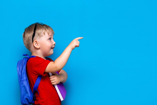 Terug naar school eerste klas junior lifestyle. kleine jongen in rode t-shirt. sluit omhoog het portret van de studiofoto van glimlachende jongen in glazen met schooltas en boek richtend met zijn vinger Premium Foto