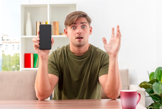 Verrast jonge blonde knappe man zit aan tafel met kopje telefoon houden