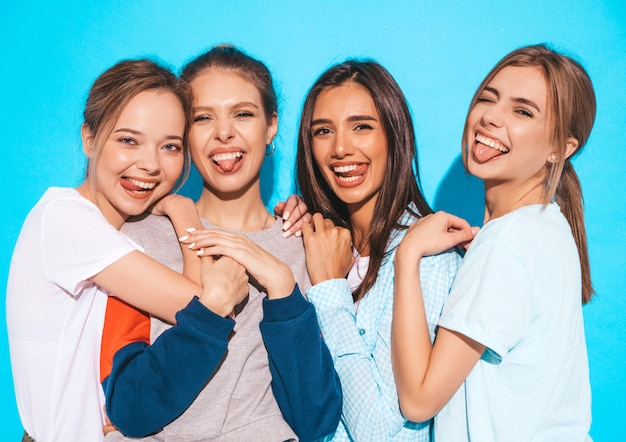 Vier jonge mooie glimlachende hipster meisjes in trendy zomerkleren. sexy onbezorgde vrouwen die dichtbij blauwe muur in studio stellen. positieve modellen die plezier hebben en knuffelen. ze tonen tongen Gratis Foto
