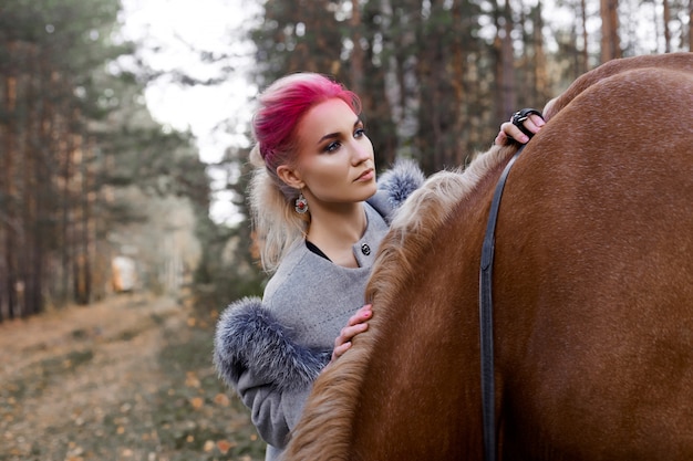 Vrouw die met de paardherfst lopen op aard. creatieve hete roze make-up meisje gezicht, haarkleuring. portret van een meisje met een paard. paardrijden in het herfstbos. herfstkleding en felrode make-up Premium Foto