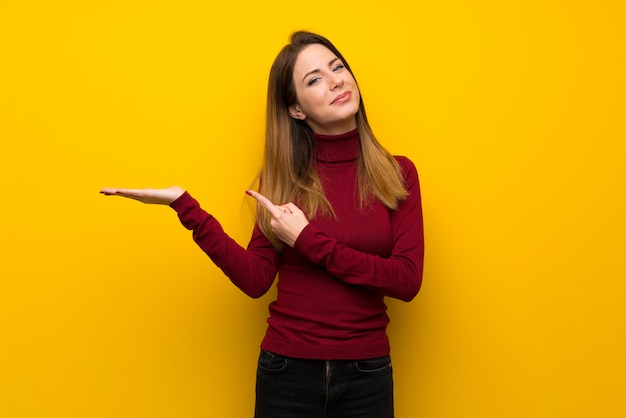Vrouw met coltrui over gele muur die copyspace denkbeeldig op de palm houdt om een advertentie in te voegen Premium Foto