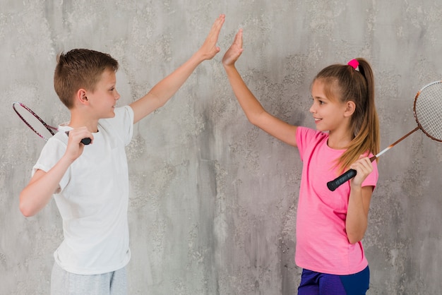 Zijaanzicht van een jongen en meisjesholding in hand racket die hoge vijf geven die zich tegen muur bevinden Gratis Foto